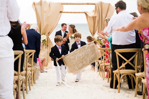 two children walk down the white sand aisle holding a sign that says "Happily Ever After" at a destination wedding on the beach in the Riviera Maya