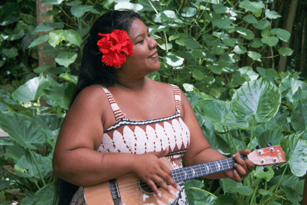 A smiling woman with a red hibiscus flower in her hair, playing a ukulele